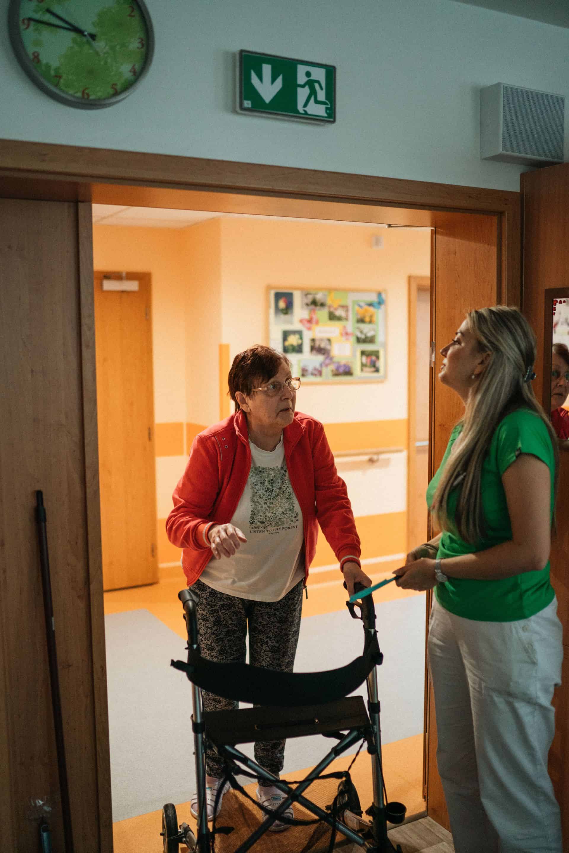 Kobieta w średnim wieku po udarze, ubrana w sportową odzież, stoi w sali rehabilitacyjnej, podpierając się na czarnym chodziku (balkoniku). Uśmiechnięta pacjentka patrzy na stojącego obok fizjoterapeutę w zielonej koszulce, który gestykuluje, wyjaśniając ćwiczenie.