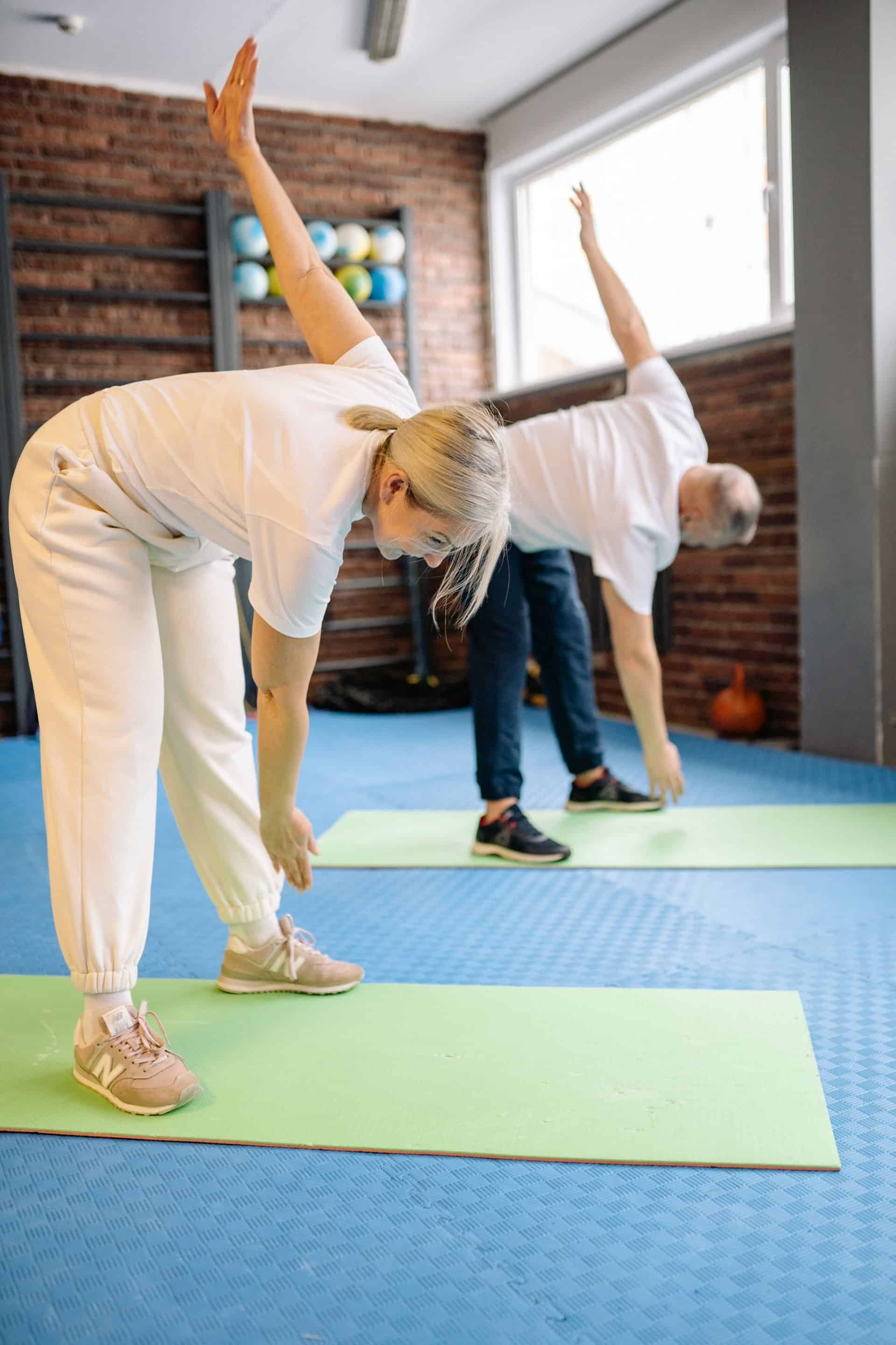 Dynamiczne ujęcie przedstawiające parę seniorów w trakcie bezpiecznego treningu usprawniającego w klinice Villa Medica. Ćwiczący w skłonie kobieta i mężczyzna wykonują wymachy rąk, co pomaga im dbać o ruchomość stawów, prawidłową koordynację oraz elastyczność kręgosłupa pod okiem wykwalifikowanego fizjoterapeuty.