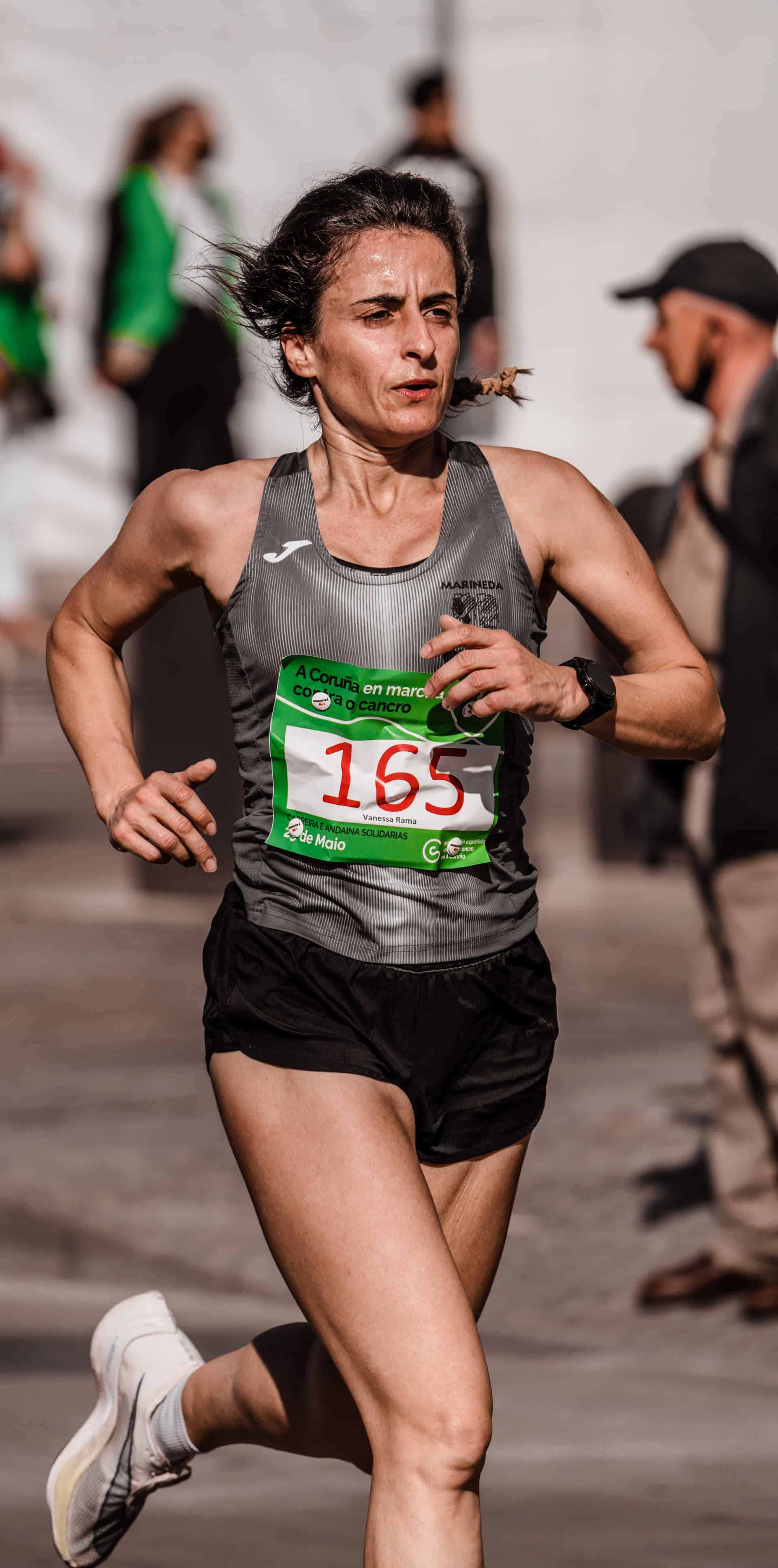 Dojrzała kobieta w stroju sportowym z numerem startowym uśmiecha się, biegnąc w maratonie po miejskiej trasie.